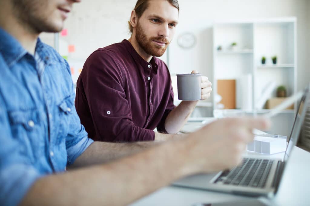 webdesigner with mug looking at laptop display while his colleague making presentation of new website