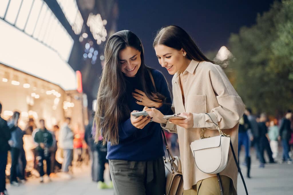 Two friends performing a "near me" search on their cell phone at night in a city.