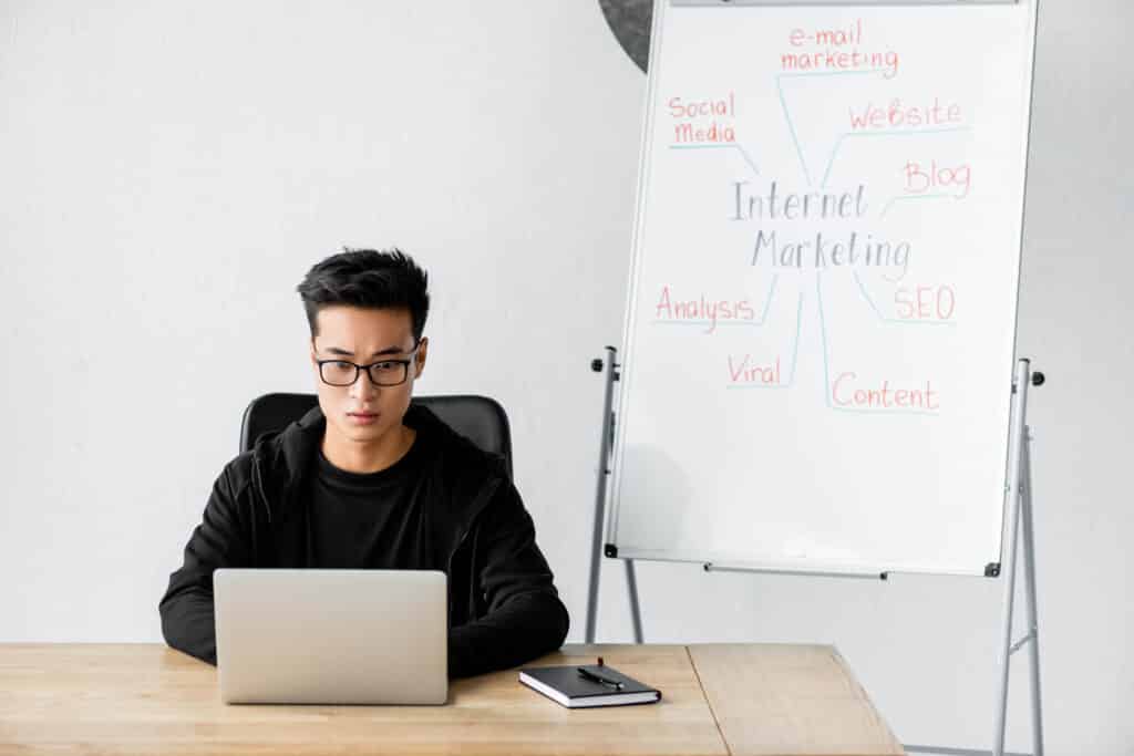 asian seo manager in glasses sitting near flipchart with lettering internet marketing on it